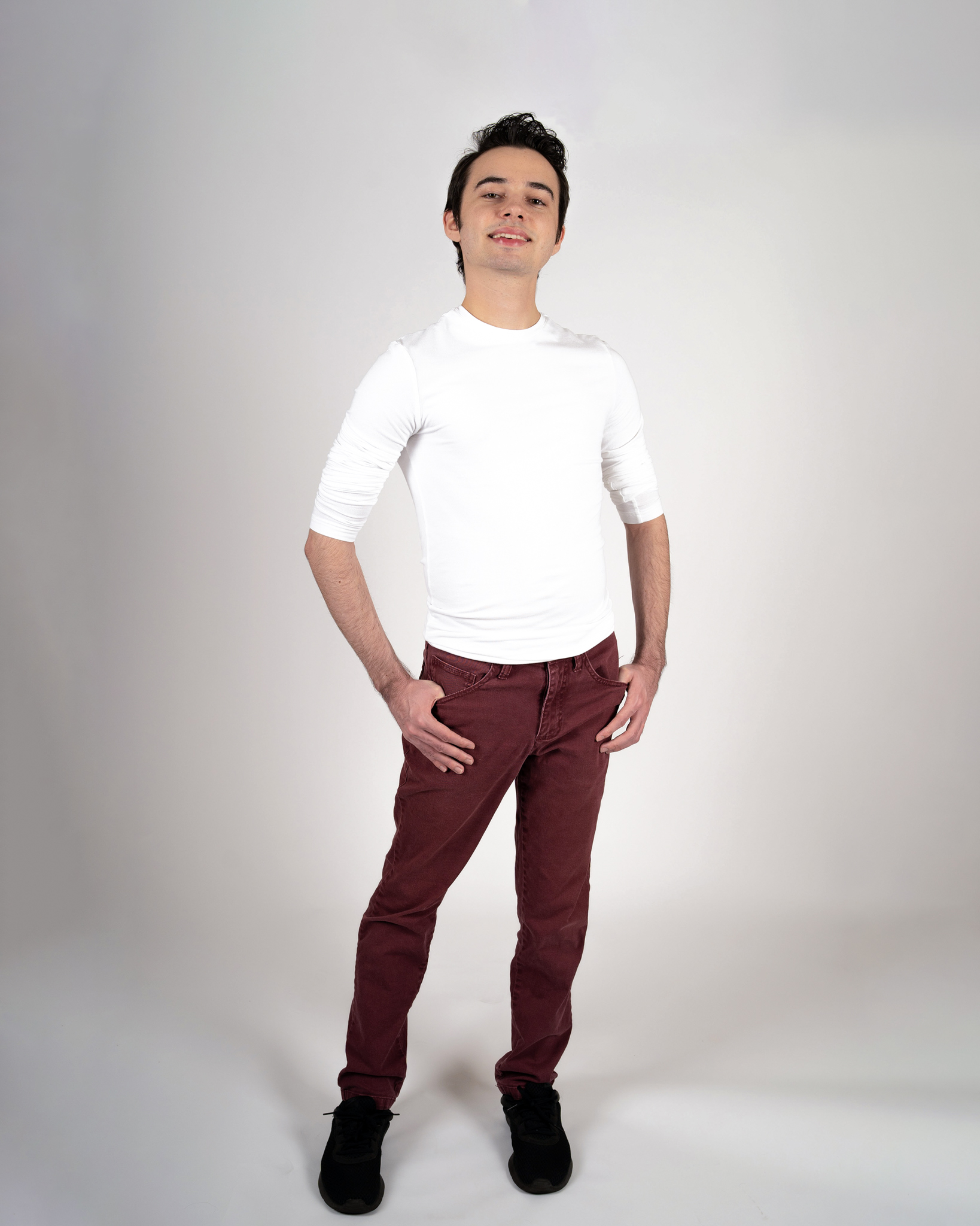 Alejandro Gonzalez, Man, posing smiling with head tilted upwards looking at the camera, in long-sleeve white shirt with rolled up sleeves, with maroon pants and black sneakers. 